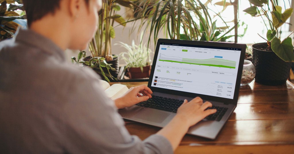 A person sitting at a wooden desk while working on a laptop with plants behind the desk