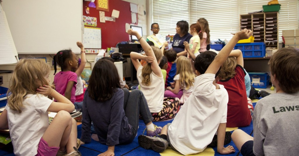 A classroom with students sitting on the floor with 3 students standing towards the front while a teacher reads a book